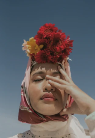 Close-up portrait of a woman wearing a pink patterned headscarf adorned with a large arrangement of red and yellow flowers against a clear blue sky