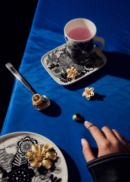 Still life photography of a cup, saucer, spoon, rings, and hand on a blue surface, creating a bright and artistic composition.