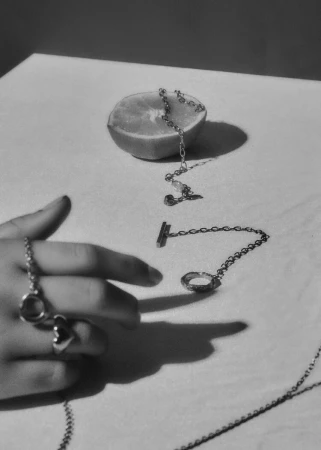 Close-up of silver jewellery chains and a half citrus fruit arranged on a flat surface with a hand resting nearby.