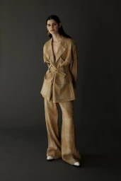 Model wearing a tan textured blazer and wide leg trousers standing against a dark grey background.