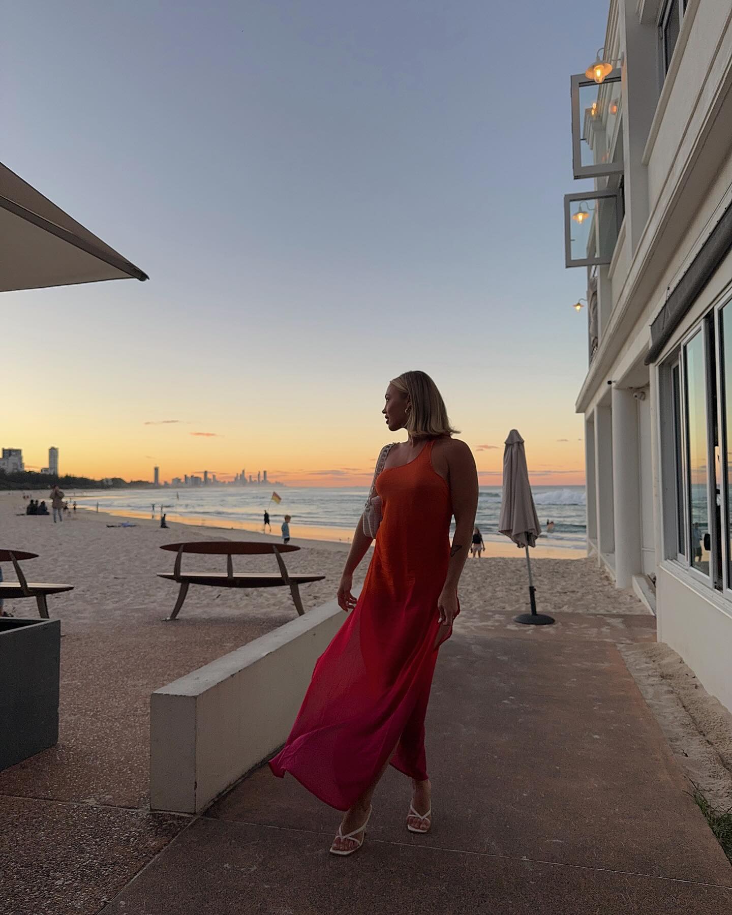 Woman in orange dress walks along a beachside path at sunset, with city skyline, ocean waves, and tables in the background.