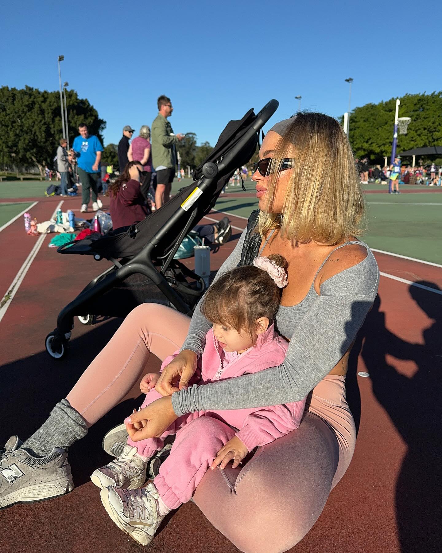 Woman and child outdoors on a sunny day near a stroller, on a sports court, with other people in the background.