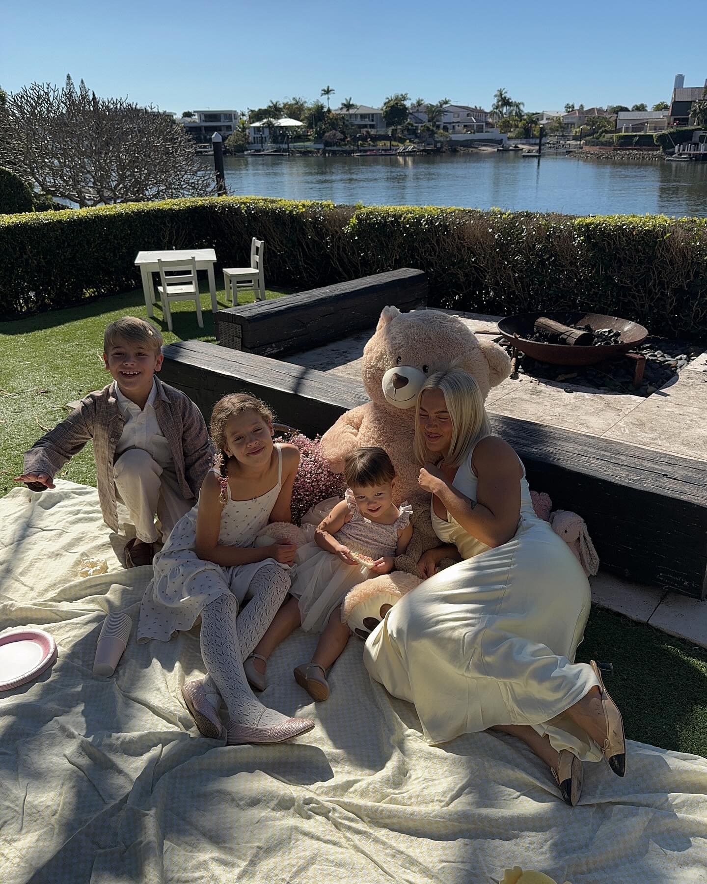 Family of five including children gathered with a teddy bear on a blanket in a yard with a waterfront view on a sunny day.