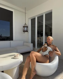Tammy Hembrow seated in a white curved armchair wearing a cut-out swimsuit on a bright balcony in Sydney