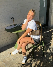Tammy Hembrow seated outdoors wearing a white t-shirt, dark skirt, and crossbody monogram handbag.