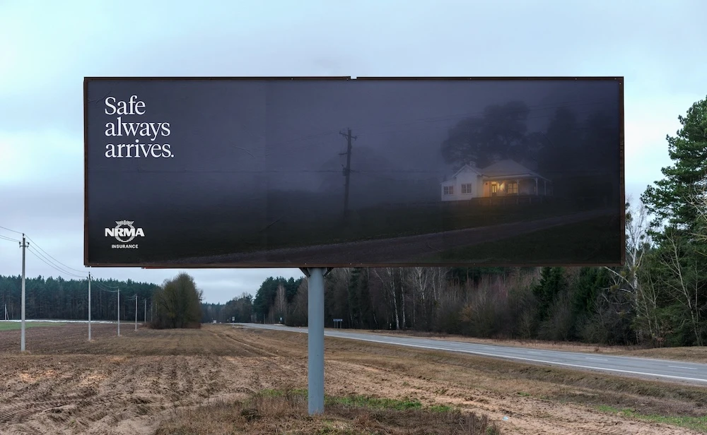 Billboard showcasing a house amidst fog at dusk, promoting NRMA Insurance with the tagline 'Safe always arrives' against a rural backdrop.