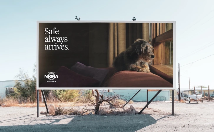 Billboard displays an image of a dog on a couch, promoting NRMA insurance, set against a desert landscape with clear blue skies.