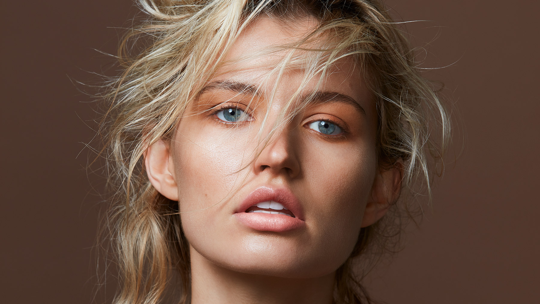Close-up portrait of a young adult female with blue eyes and blonde hair against a brown background in a photo studio setting.