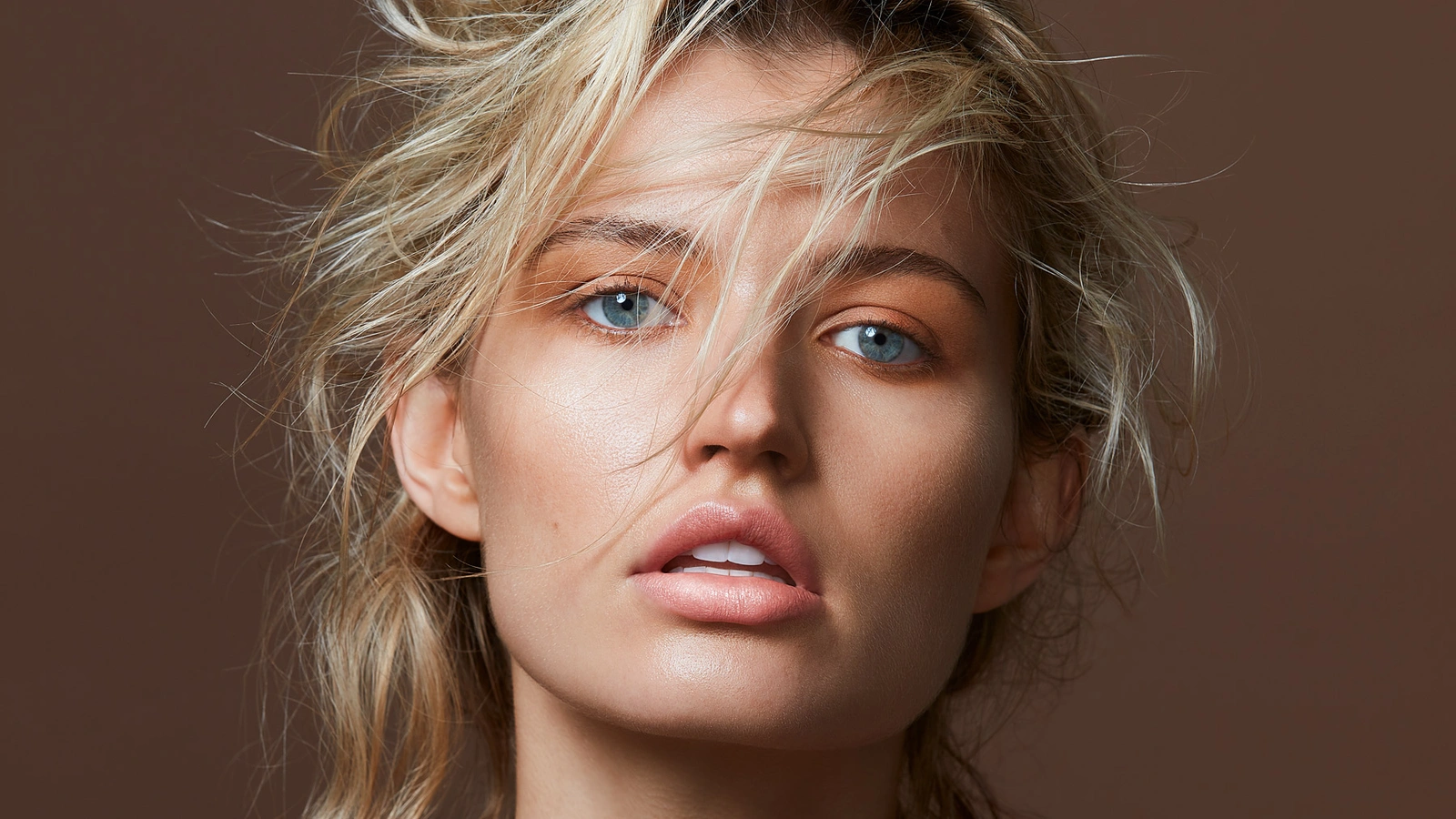 Close-up portrait of a woman with tousled blonde hair and blue eyes against a solid brown background, retouched by Jamie Phillips.