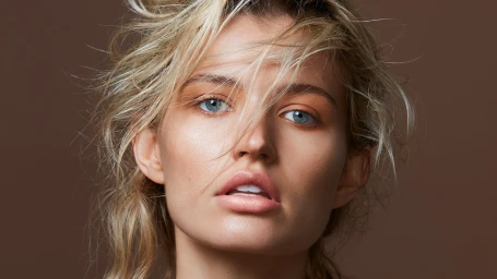 Close-up portrait of a woman with tousled blonde hair and blue eyes against a solid brown background, retouched by Jamie Phillips.