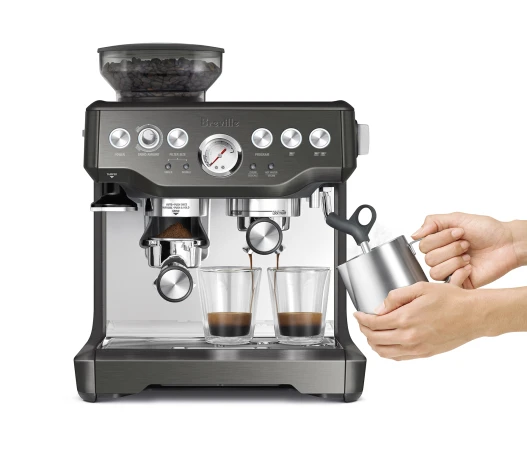 Espresso machine on a white surface, pouring espresso into two clear glass cups, with a person steaming milk in a metal pitcher.