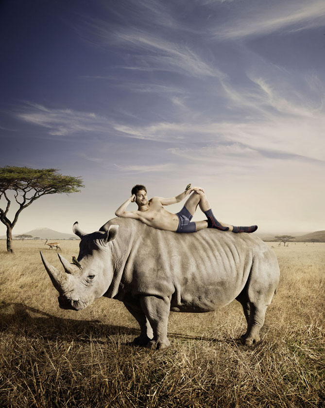 Man reclines on a rhinoceros in an African savanna-style environment under a blue sky with wispy clouds and a lone acacia tree.
