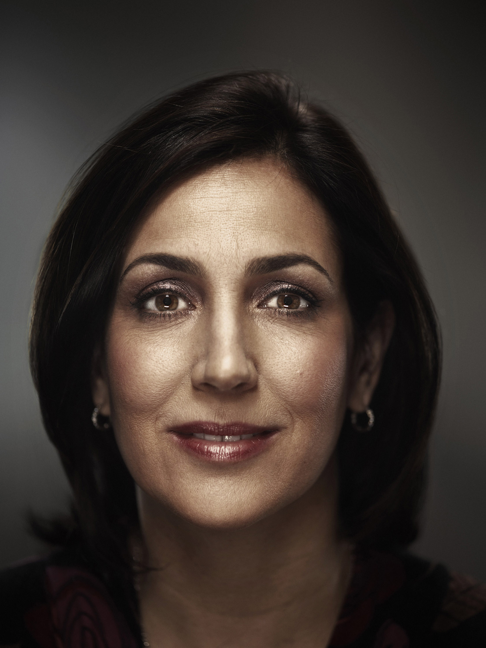 Close-up portrait of a mature adult woman with brown hair and eyes, wearing earrings, against a dark gray background.