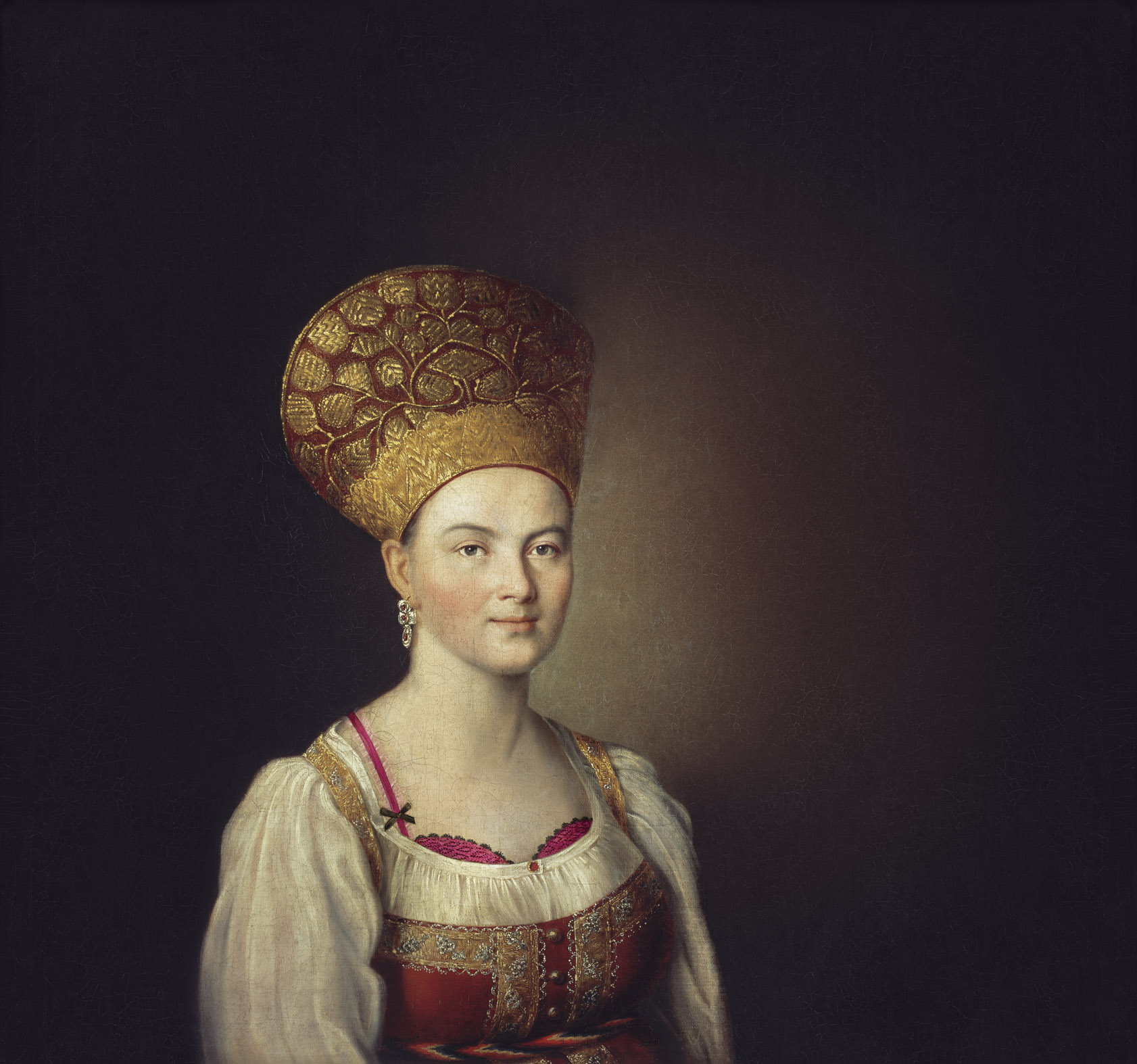 Portrait of a woman wearing an elaborate gold headdress and a red and gold dress, set against a dark brown background.