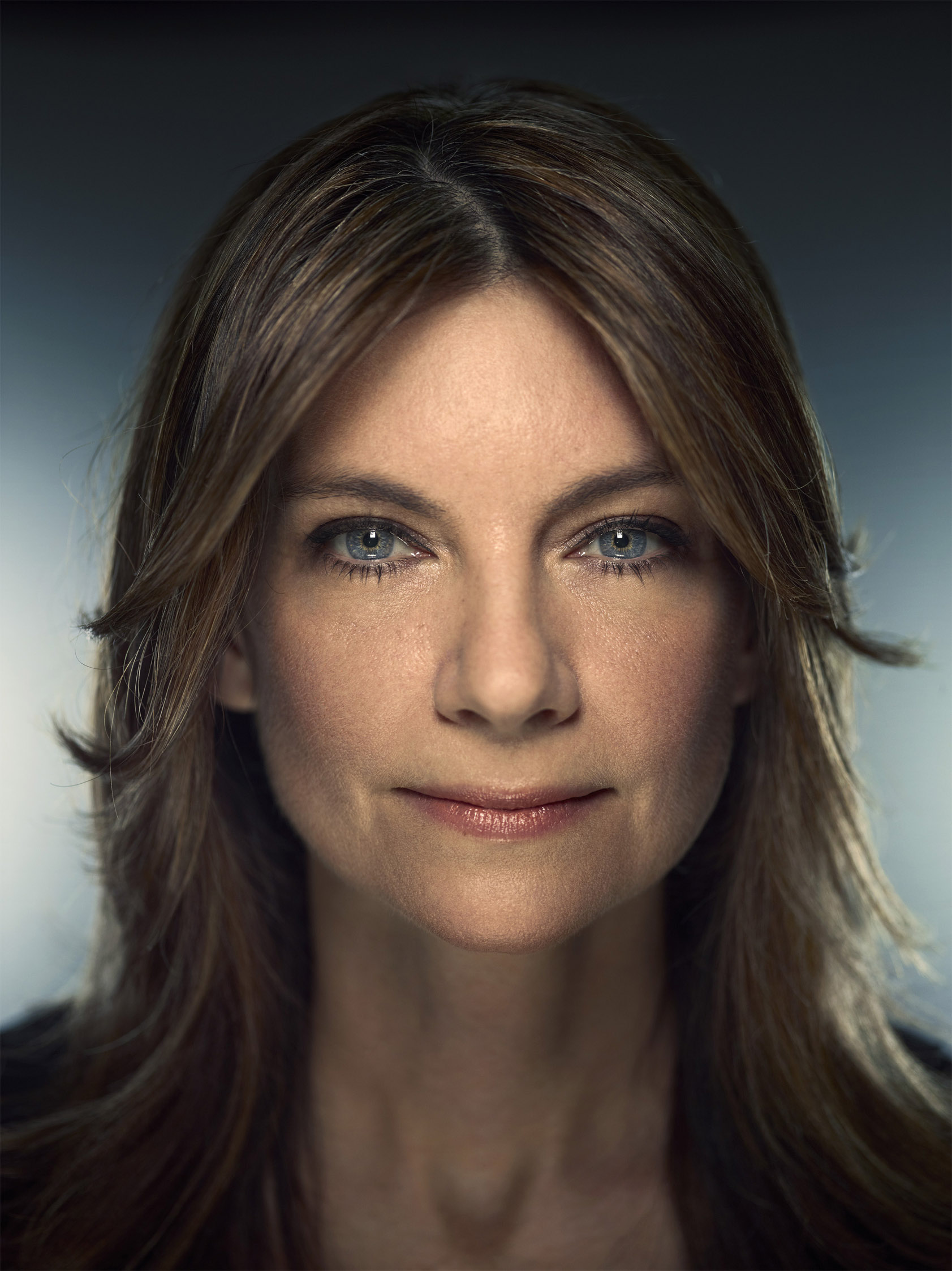 Close-up portrait of a mature woman with medium-length brown hair, fair skin, and light blue eyes, gazing directly at the camera in a studio setting.