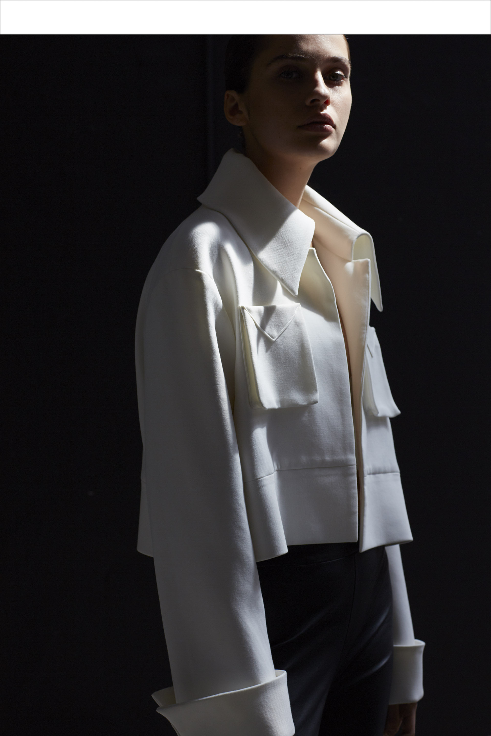 Fashion shot of a woman wearing a white jacket in a dark studio. The jacket has large collar and pockets. Dramatic lighting and high contrast.