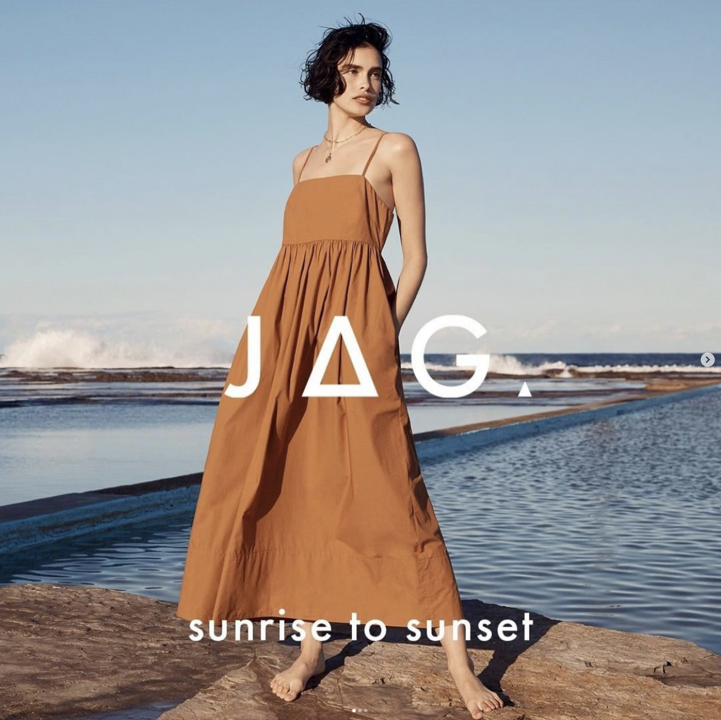 Brunette woman in a sleeveless rust-colored dress posing barefoot on rocks by a pool with an ocean view on a sunny day.