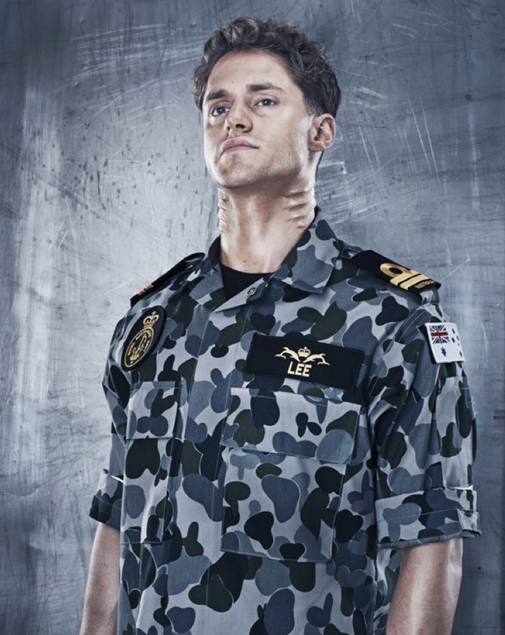Portrait of a young adult male wearing a camouflage-patterned military uniform, looking upwards against a grey industrial background.