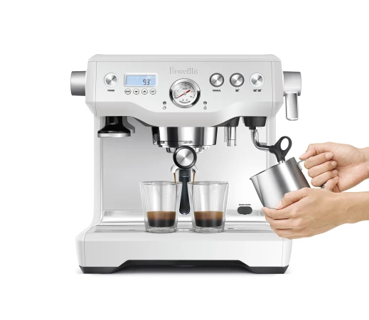 White Breville espresso machine pouring espresso into two clear glasses with a woman's hands holding a metal milk steaming pitcher.