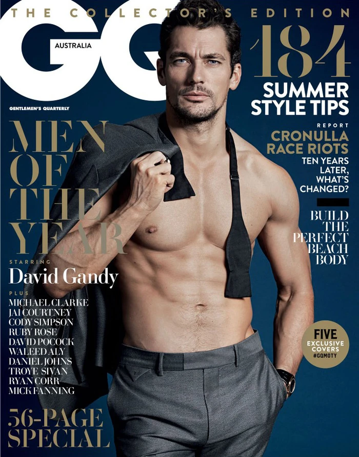 Shirtless male model David Gandy holding a grey suit jacket over his shoulder against a dark background