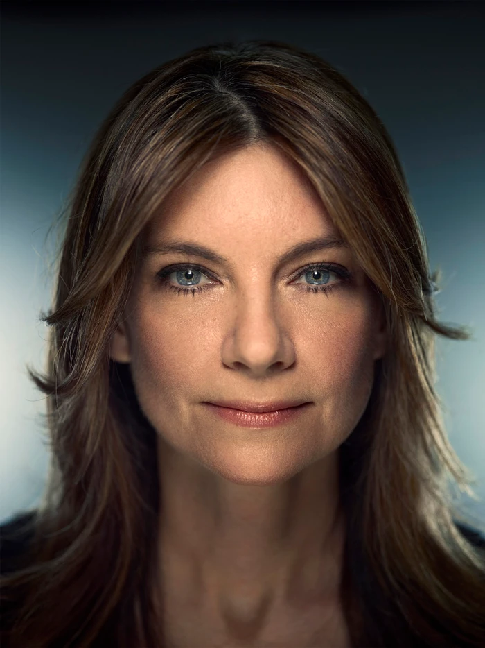 Close-up portrait of a woman with long brown hair and blue eyes, looking directly at the camera with a subtle expression.