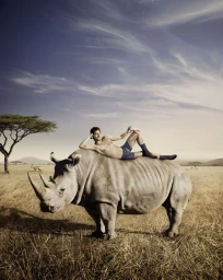 A man wearing blue underwear and patterned socks lounging on the back of a large rhinoceros in a dry savannah landscape.