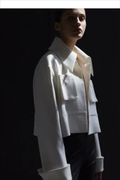 A portrait of a model wearing a structured white jacket against a dark background, retouched by Judith Caingat.