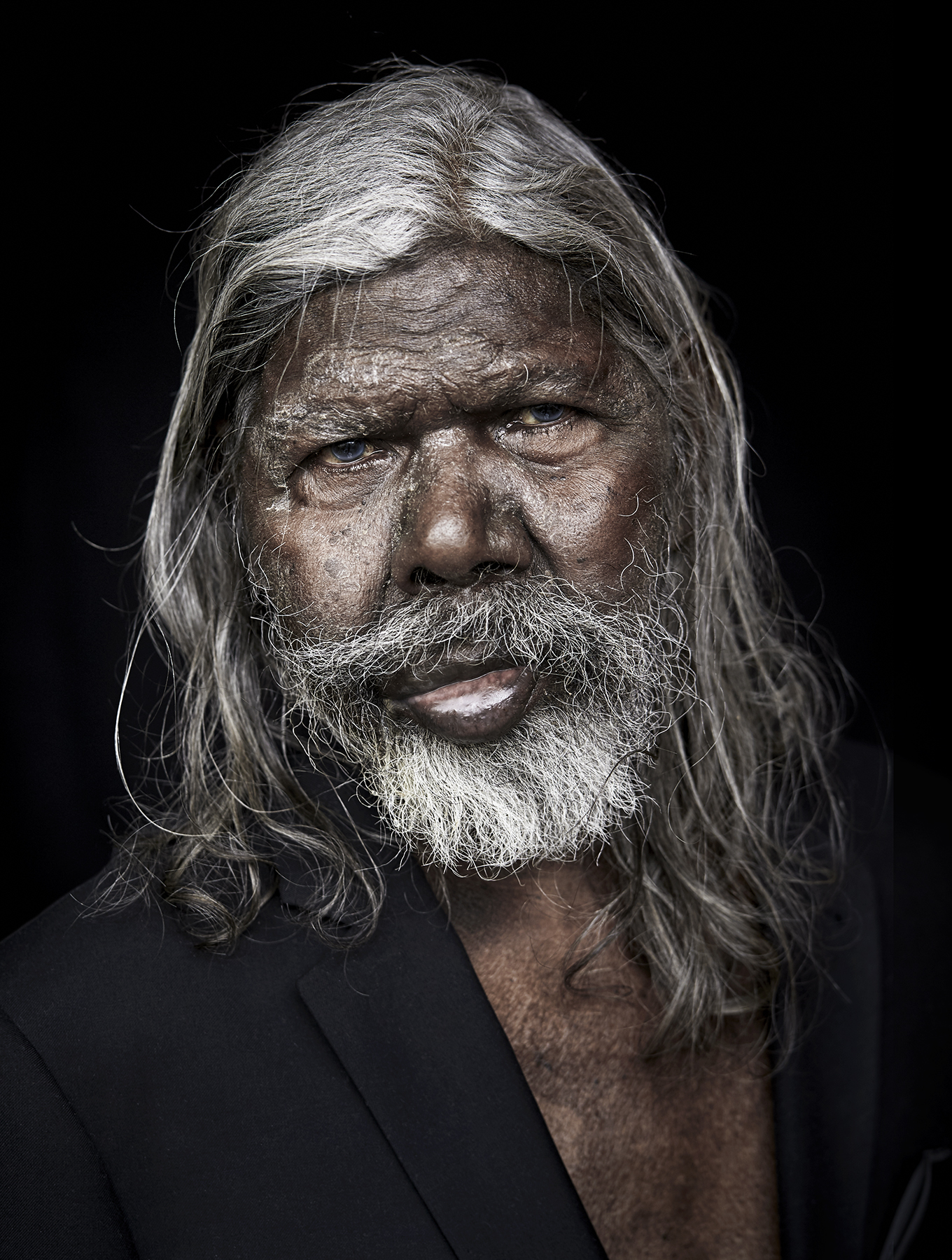 Portrait of an elderly Aboriginal man with white hair and beard, wearing a dark jacket against a stark black background.