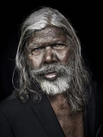 Portrait of an elderly Aboriginal man with white hair and beard, wearing a dark jacket against a stark black background.