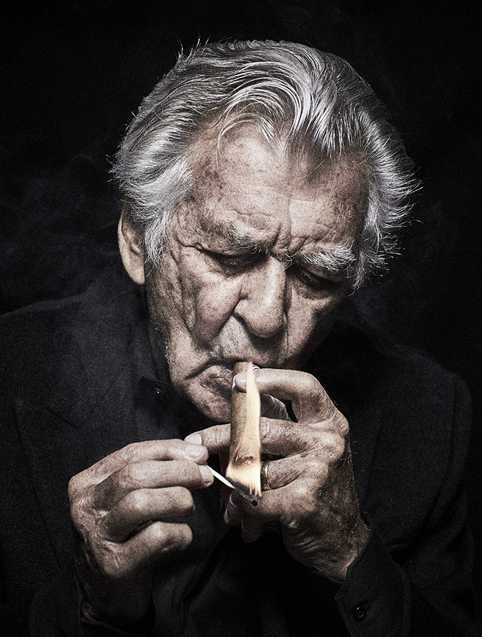 Portrait of an older man lighting a cigar with a match in a dark, indoor setting, showing details of his face, hands, and clothing.
