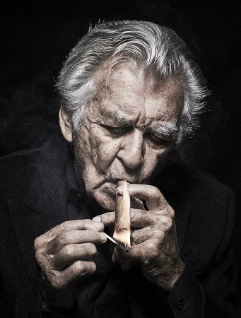 Portrait of an older man lighting a cigar with a match in a dark, indoor setting, showing details of his face, hands, and clothing.