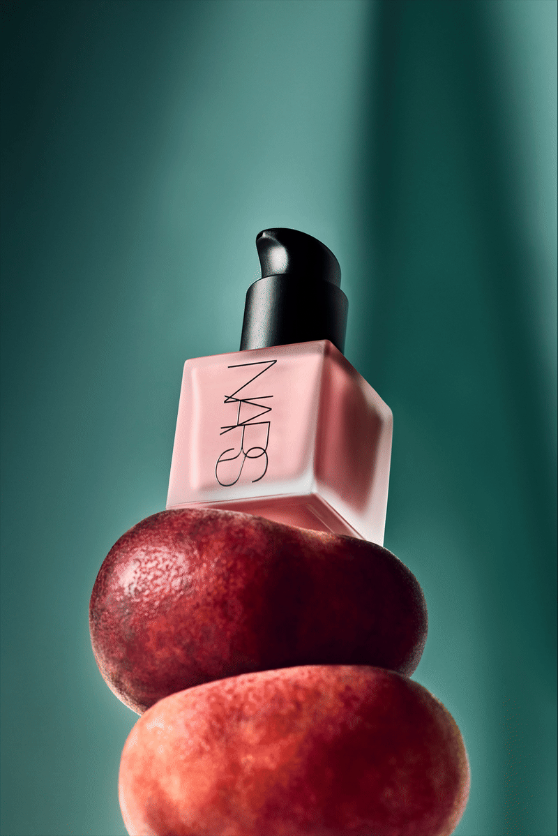 NARS cosmetic bottle on stacked peaches against teal backdrop. Product shot showcases beauty and healthy skincare concept.