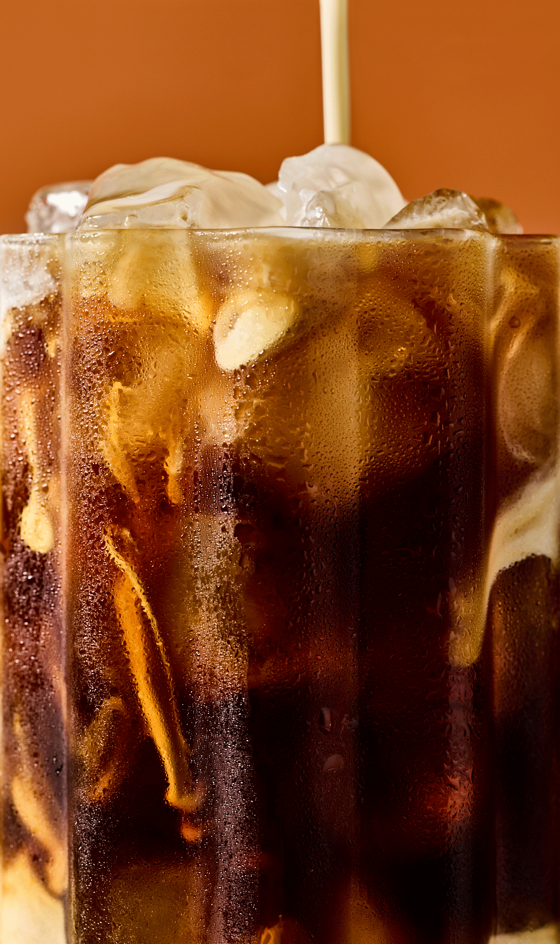 Close-up of iced coffee drink in tall glass as cream is being poured in. Refreshing beverage with ice cubes and condensation.