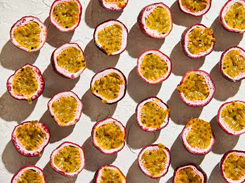 Overhead shot of sliced passion fruit halves with yellow pulp and black seeds arranged on a textured light beige surface.