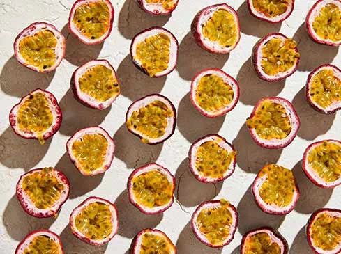 Overhead shot of sliced passion fruit halves with yellow pulp and black seeds arranged on a textured light beige surface.