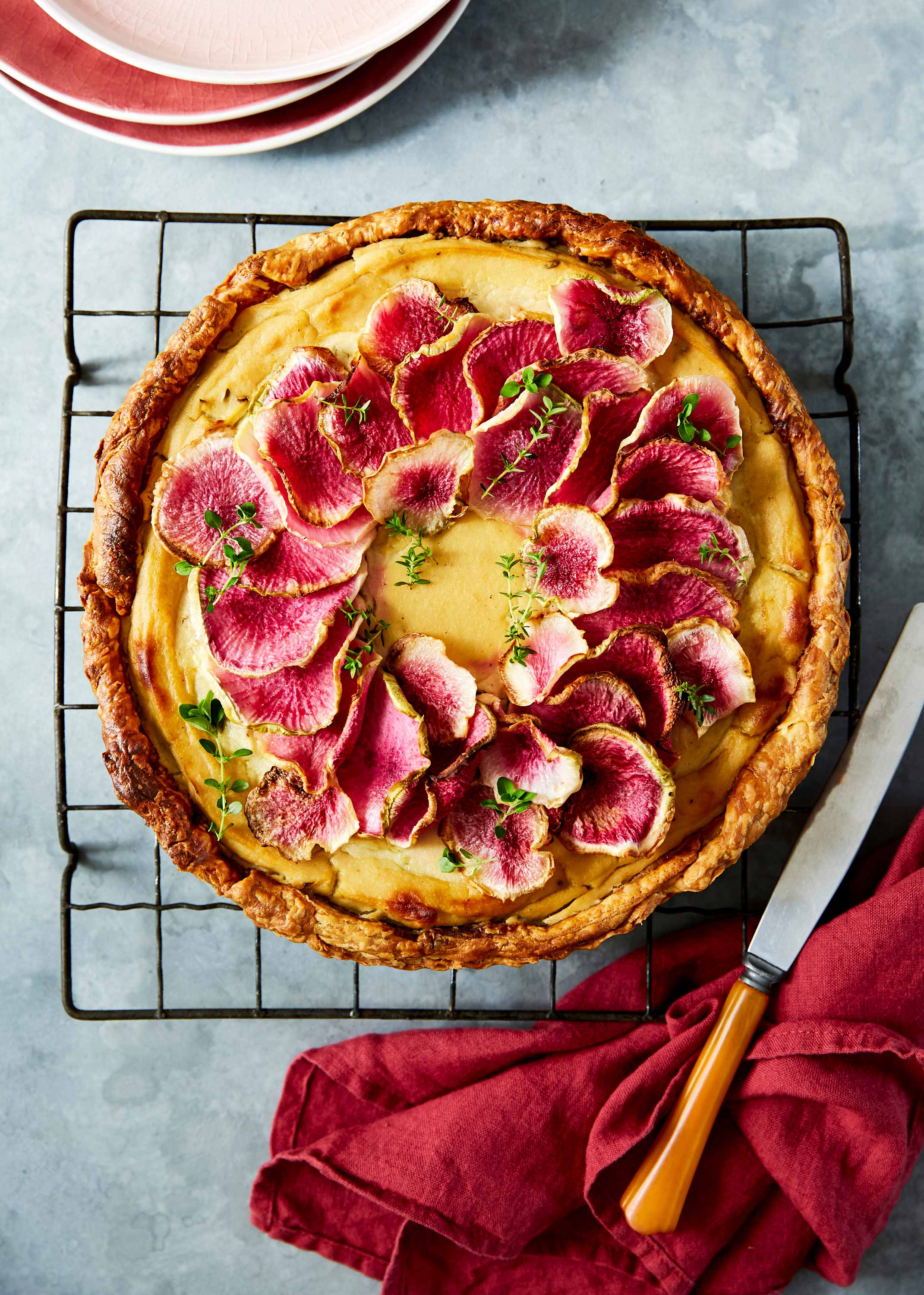 Overhead shot of a radish tart with golden crust, topped with radish slices and herbs, displayed on a black cooling rack with red linen.