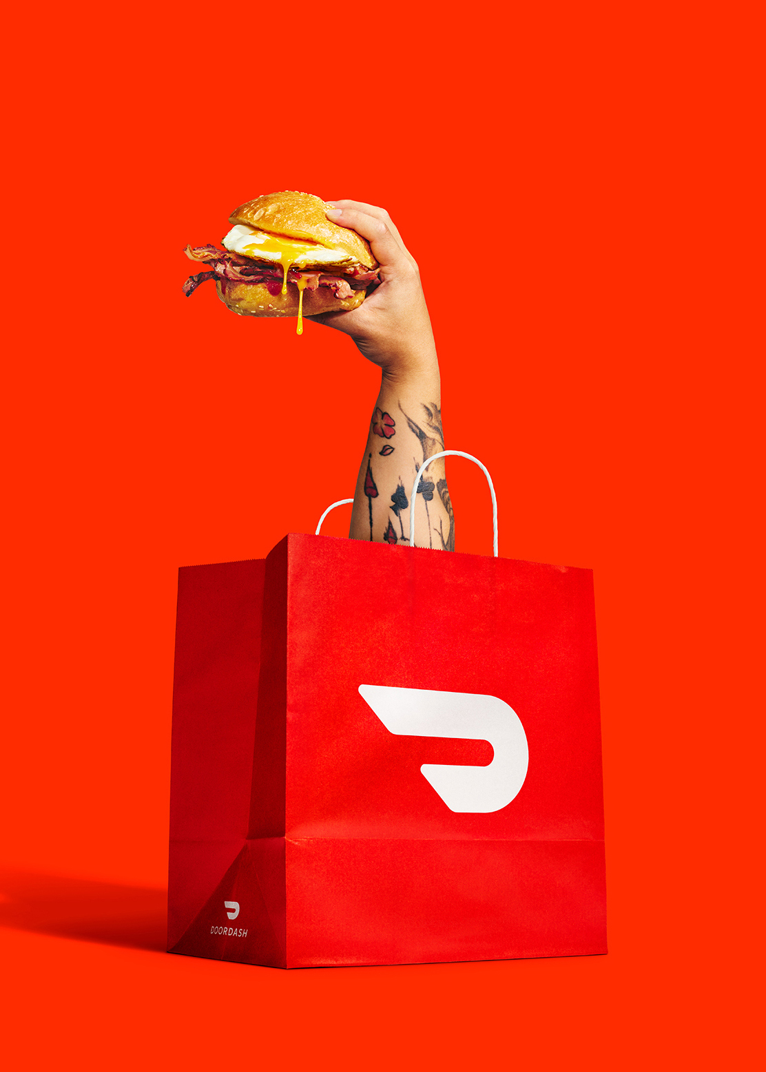 Hand holding a sandwich emerging from a red DoorDash take-out bag, set against a bright red background. Food and delivery concept.