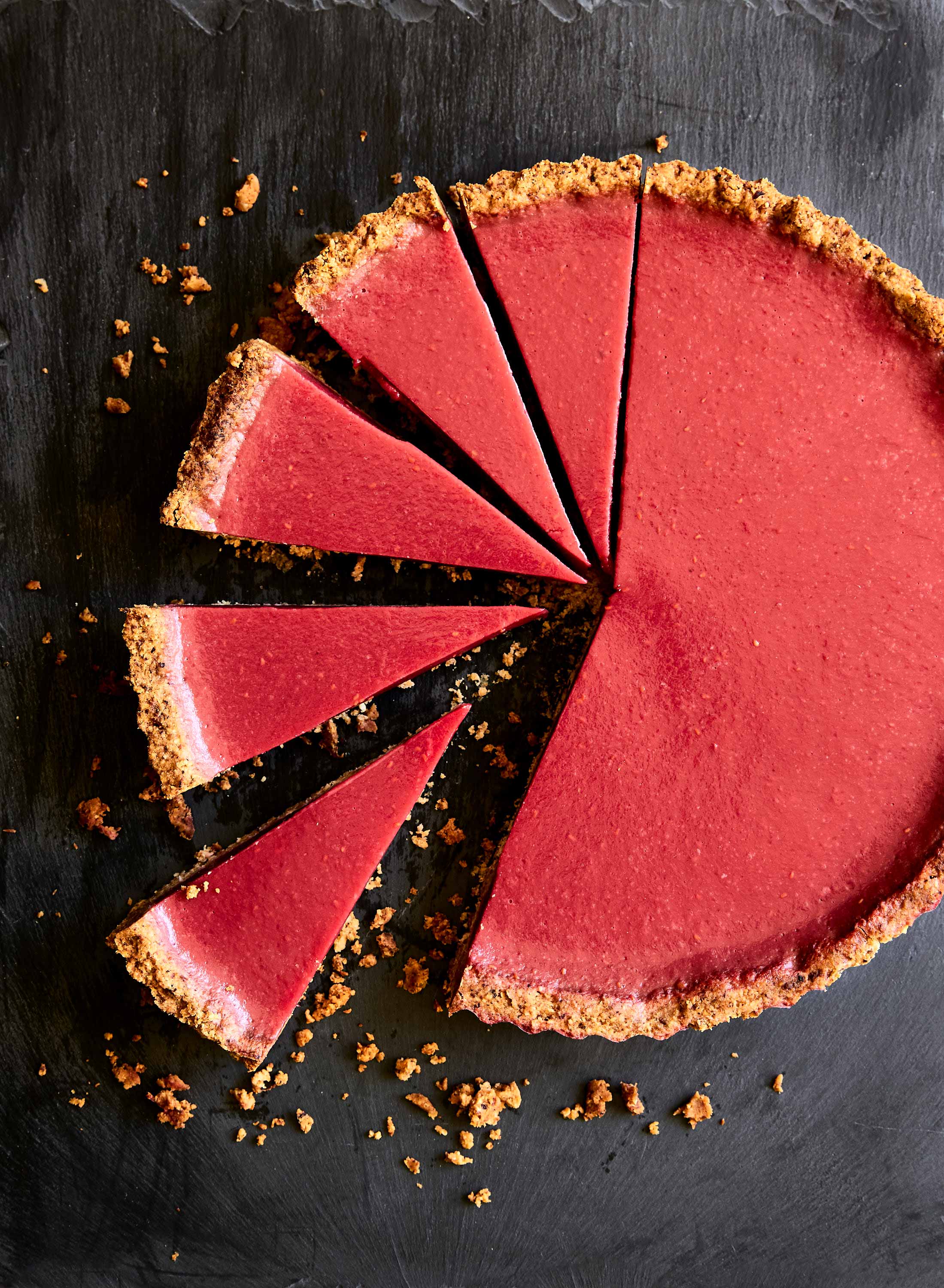 Overhead shot of a vibrant pink tart with a graham cracker crust, cut into slices and arranged on a dark background, showcasing a delightful dessert.