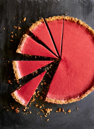Overhead shot of a vibrant pink tart with a graham cracker crust, cut into slices and arranged on a dark background, showcasing a delightful dessert.