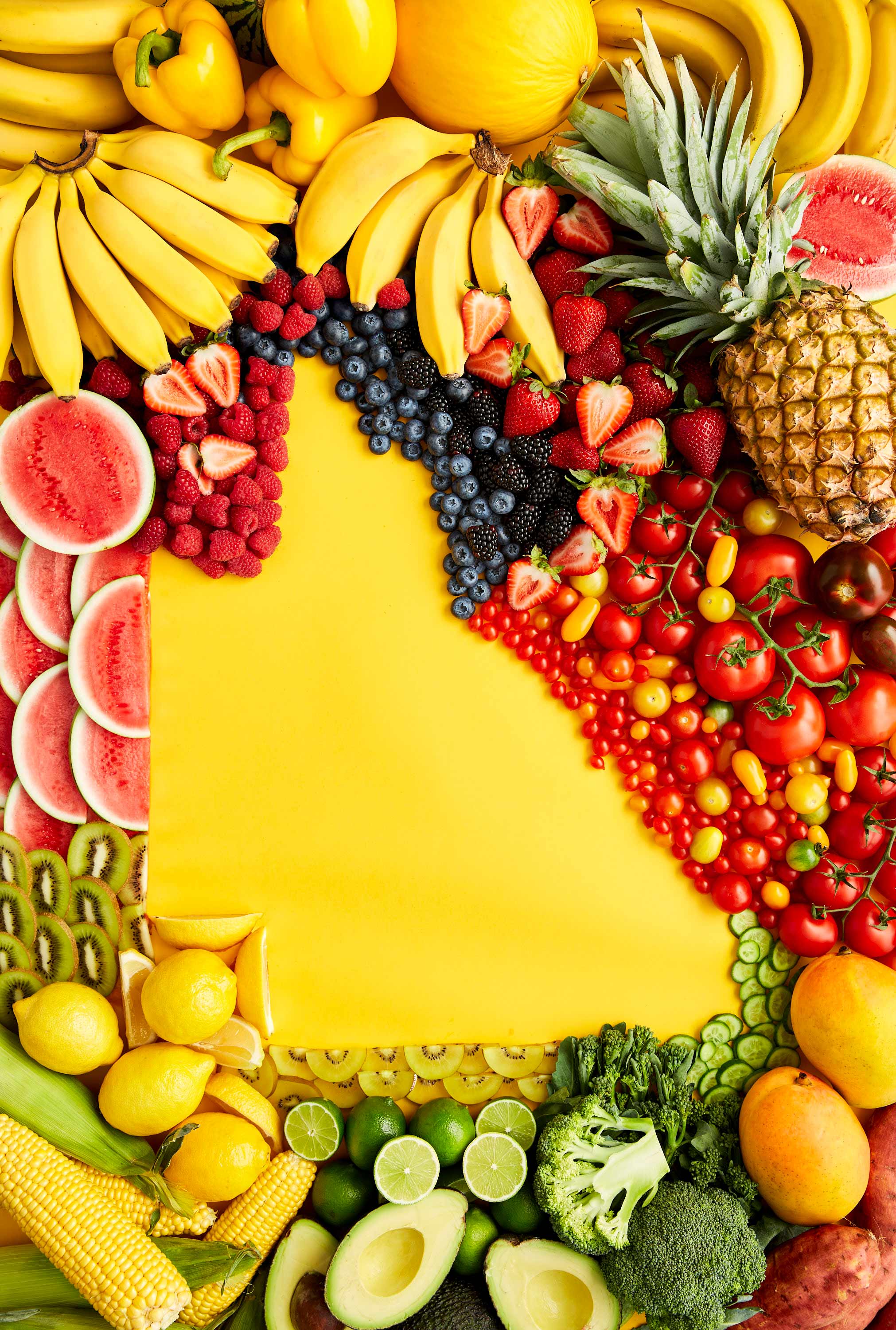 Overhead shot of assorted fresh fruits and vegetables arranged around a bright yellow backdrop, showcasing a colorful variety of produce.