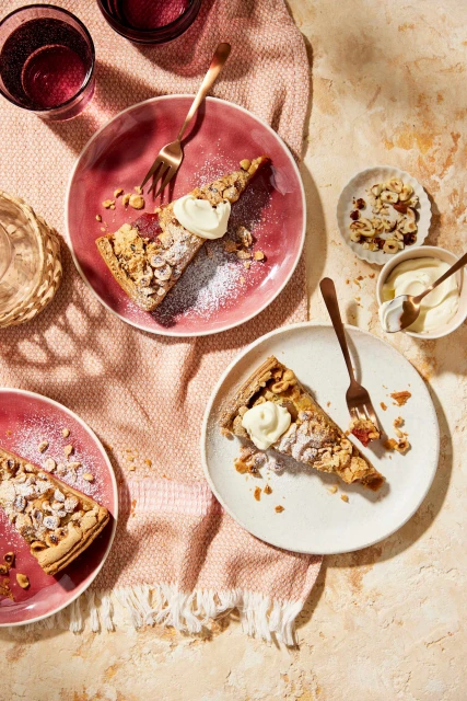 Overhead shot of three plates of hazelnut tart slices with forks, set on a pink fabric and a textured beige surface, warm lighting.