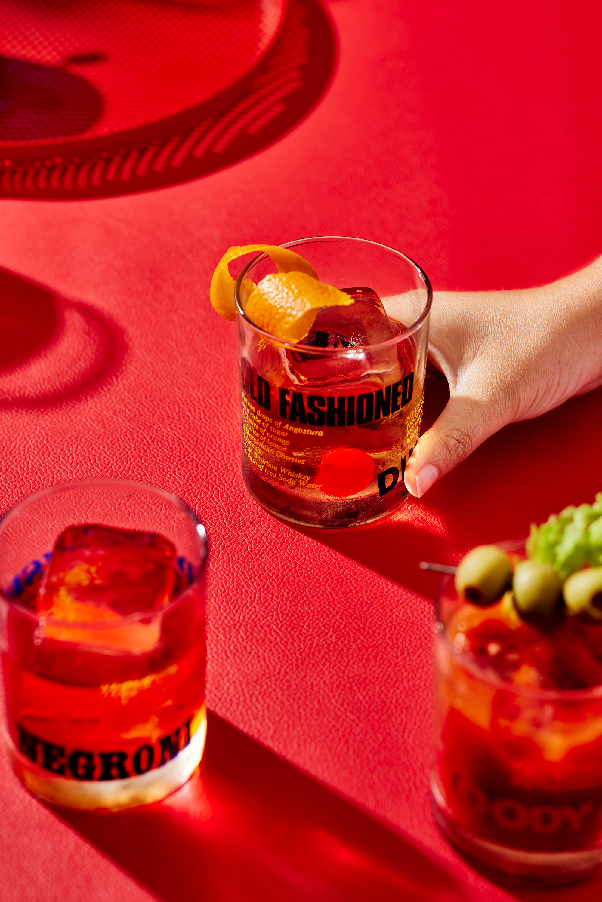 Three cocktail glasses, one held by a hand, rest on a vibrant red surface. Garnished drinks feature ice cubes, orange peels, and olives.