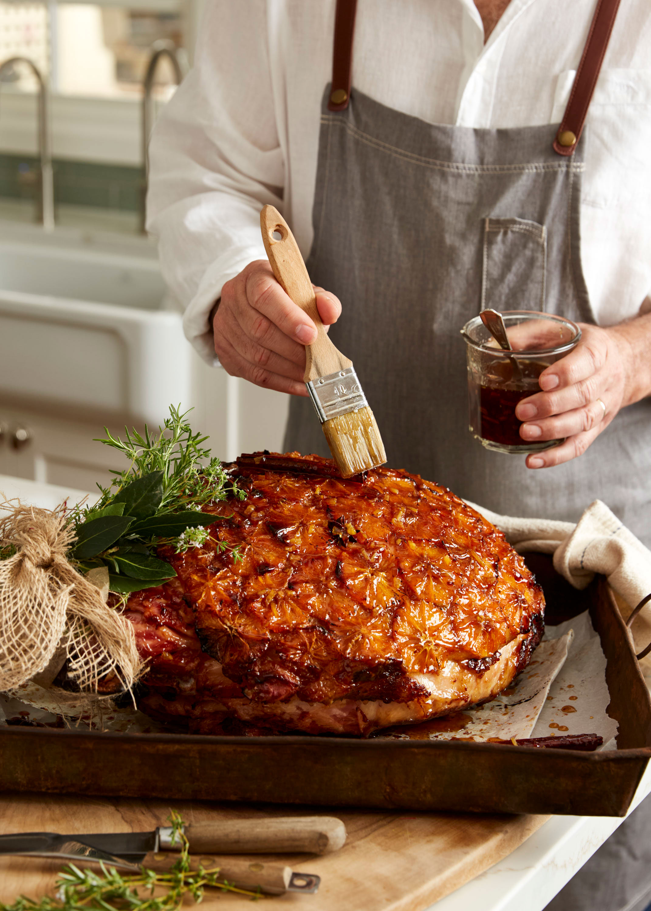 Close-up of glazed ham on a tray being brushed with sauce, adorned with herbs, in a kitchen, with a person wearing an apron.