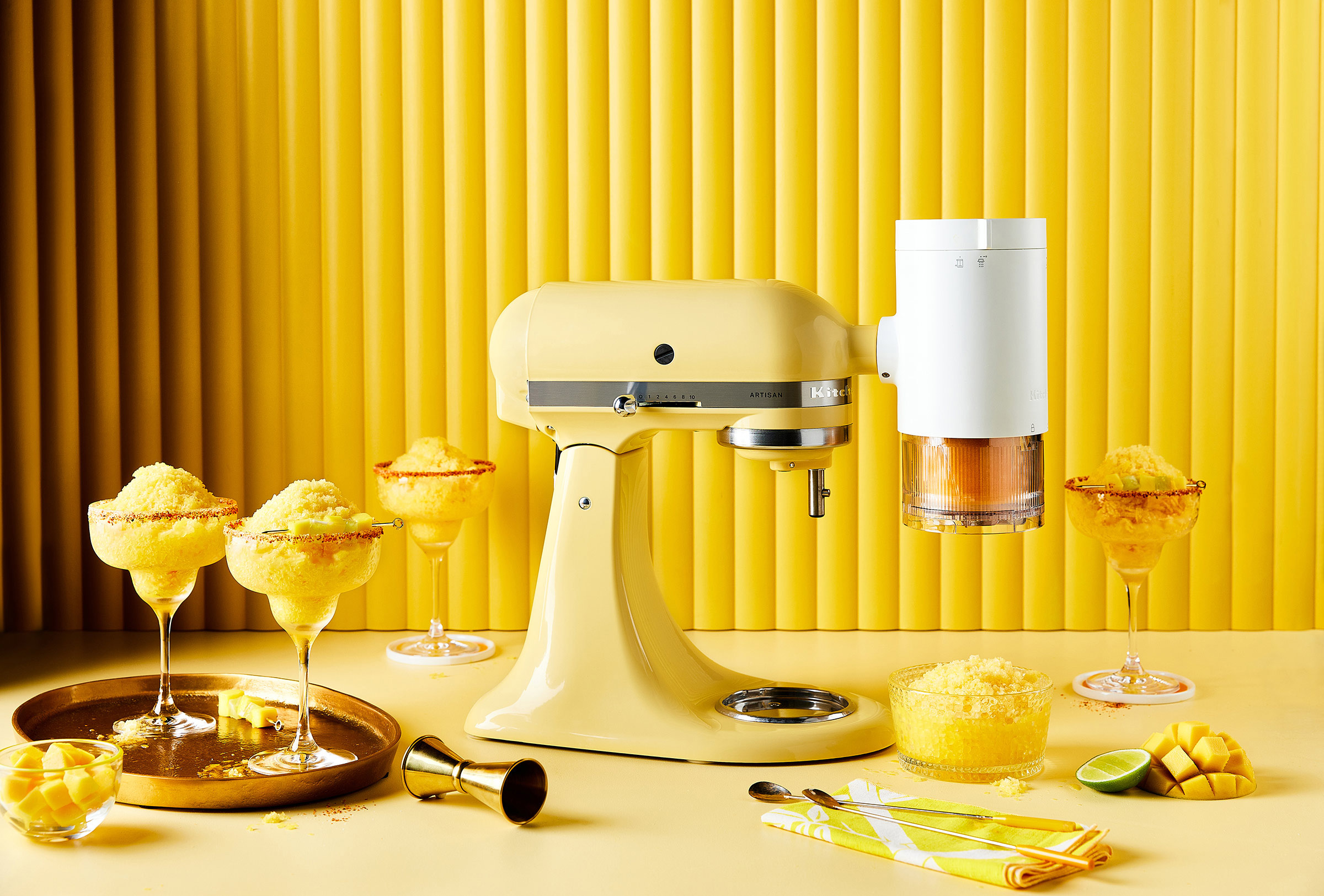 Yellow KitchenAid stand mixer with citrus juicer attachment and cocktails, set against a bright yellow background and tabletop.