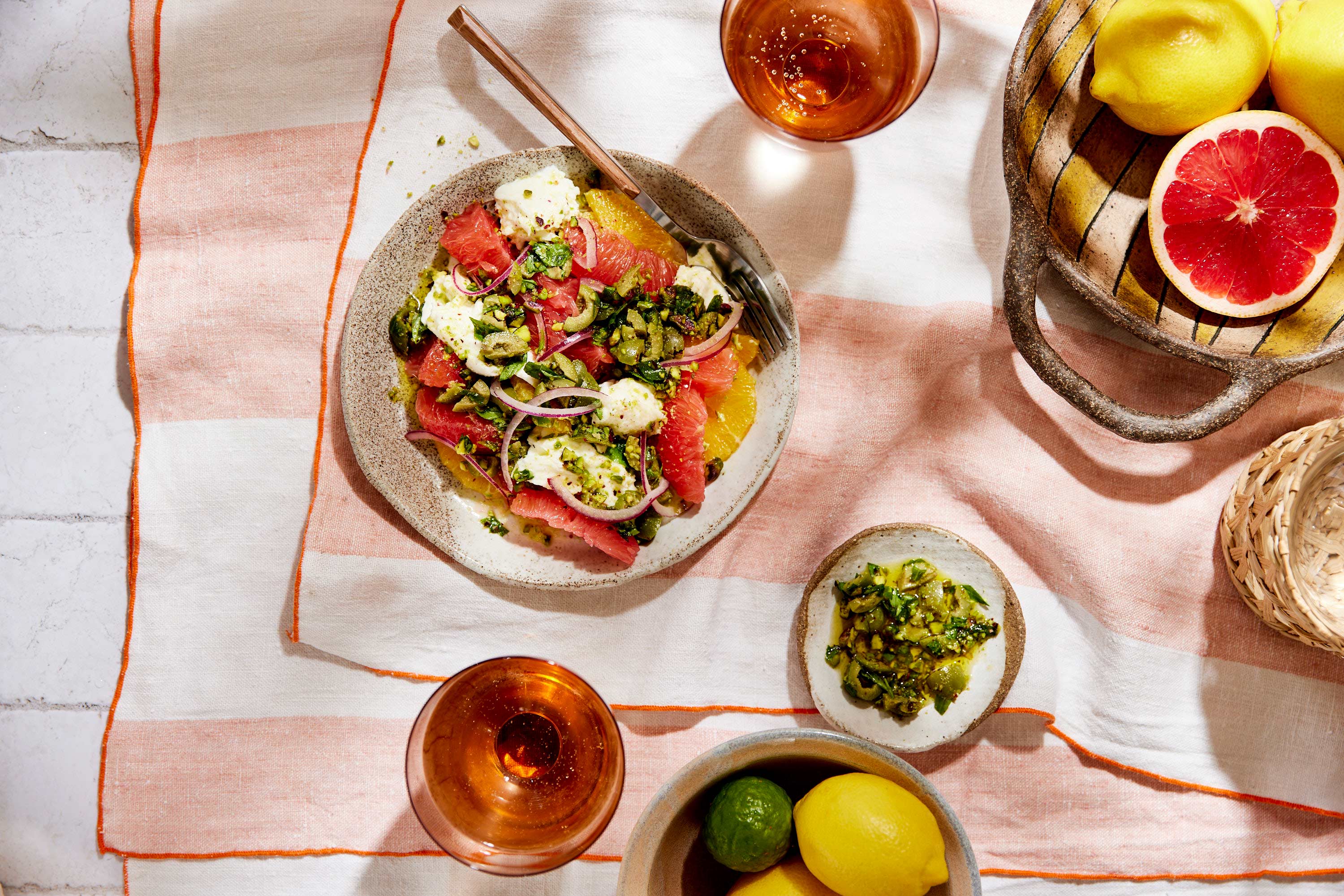 Overhead shot of citrus salad with grapefruit, orange, mozzarella, and herbs, served on a ceramic plate, with fruit and cocktails.
