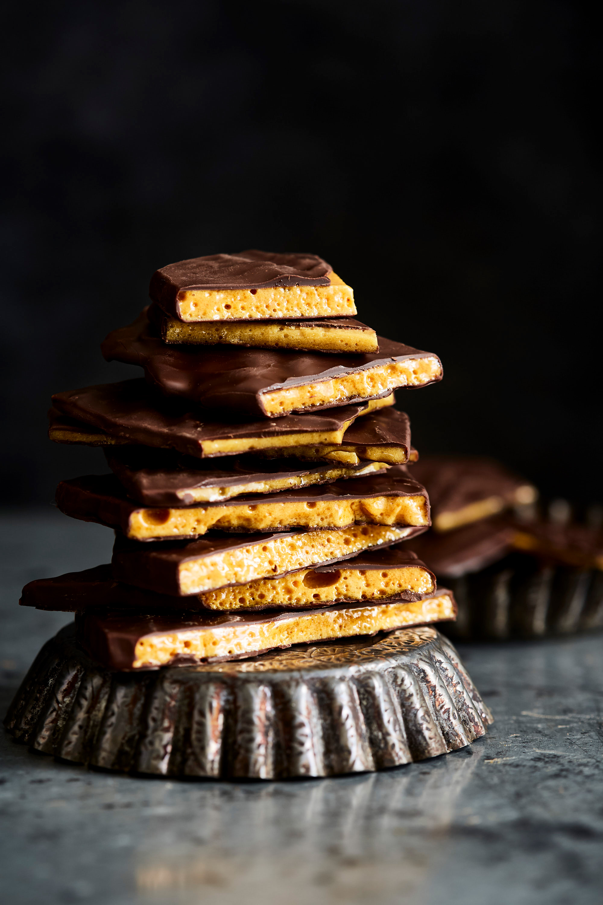 A stack of chocolate-covered honeycomb candy pieces on a metal pedestal, with another pedestal in the background, against a dark backdrop.