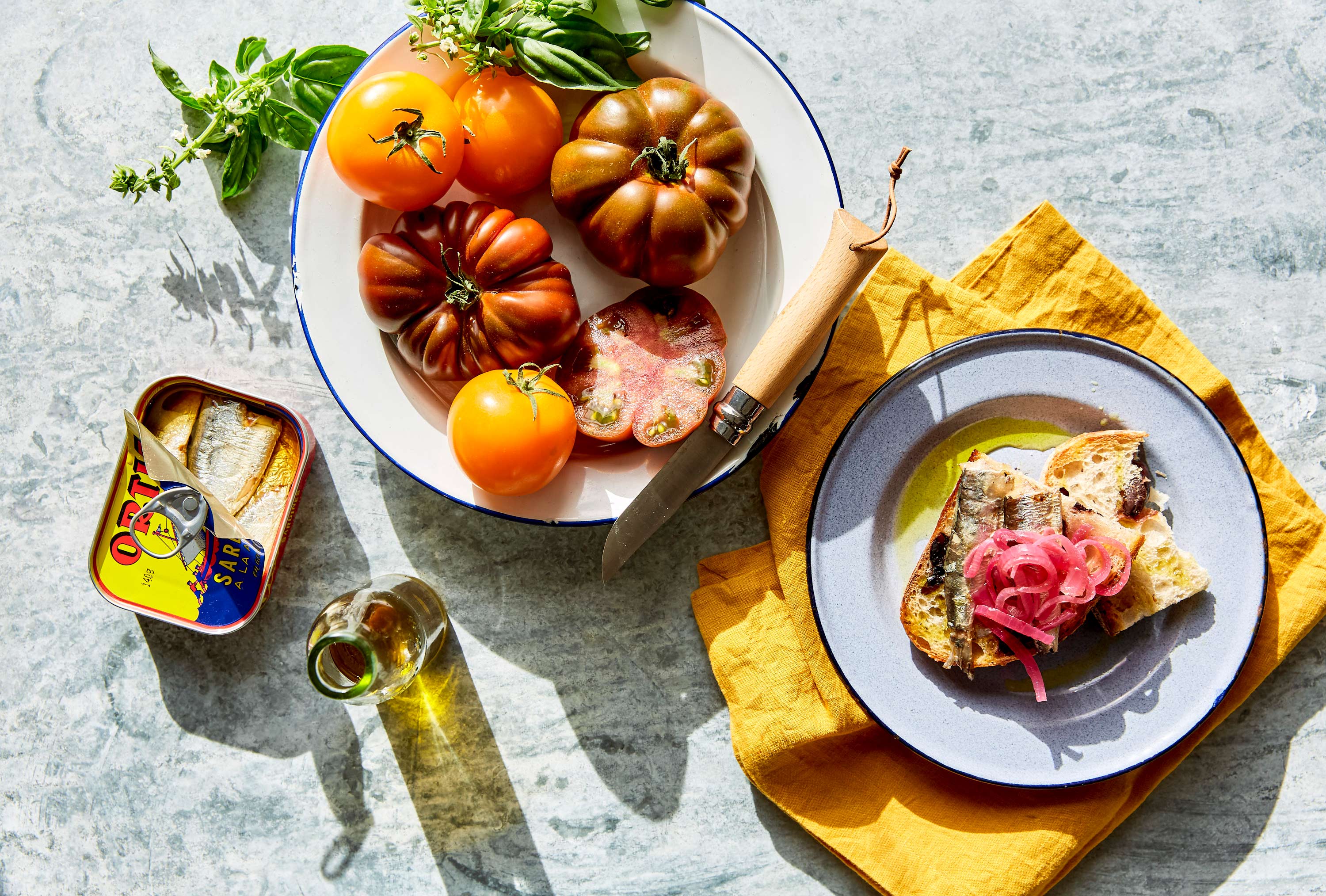 Overhead food shot of rustic heirloom tomatoes, bread with sardines and pickled onions, olive oil, and basil leaves on a table.