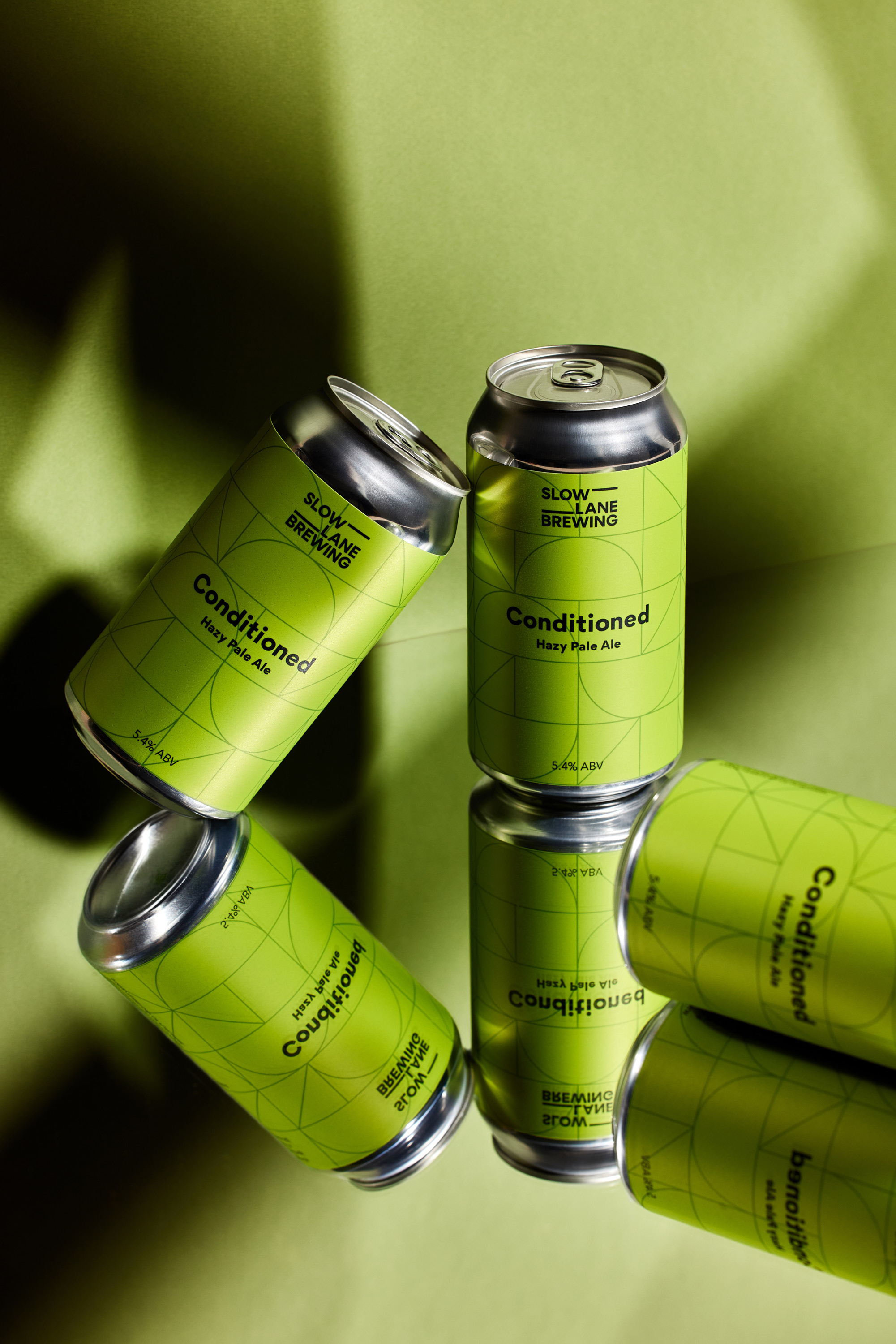 Elevated shot of three cans of Slow Lane Brewing Conditioned Hazy Pale Ale with geometric decoration against a green background and mirrored surface.