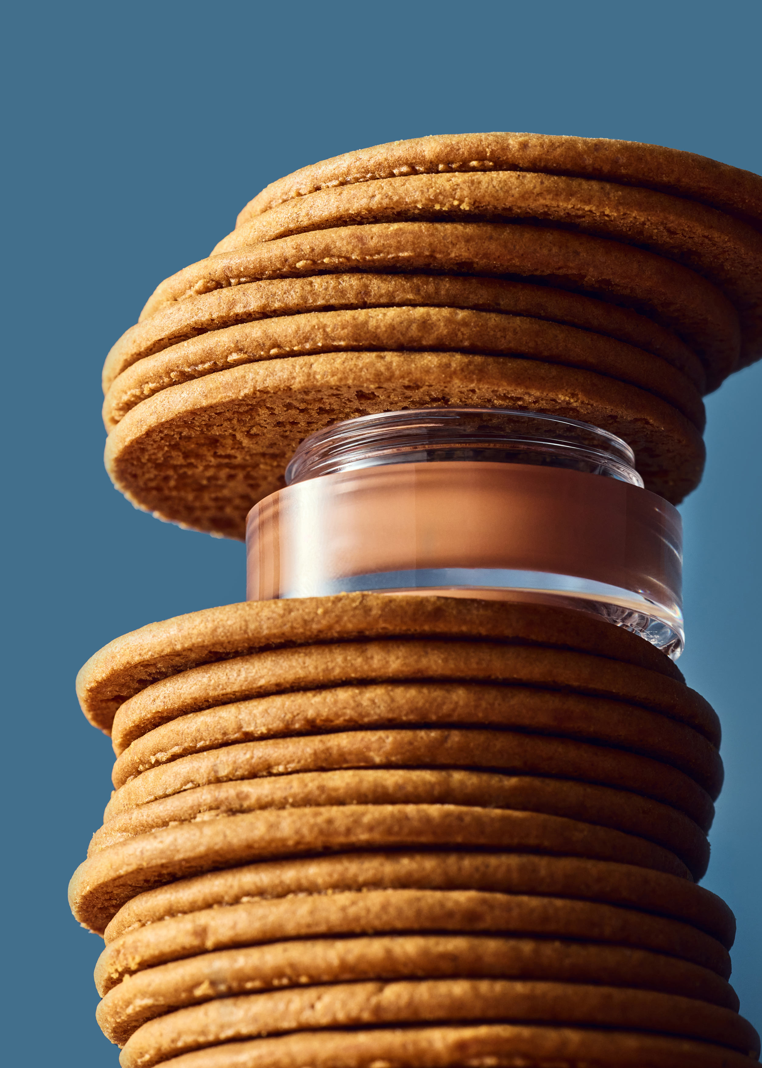 Elevated perspective of round cookies stacked high with beige foundation jar in between, set against a blue backdrop.