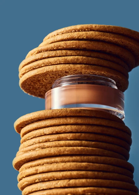 Elevated perspective of round cookies stacked high with beige foundation jar in between, set against a blue backdrop.